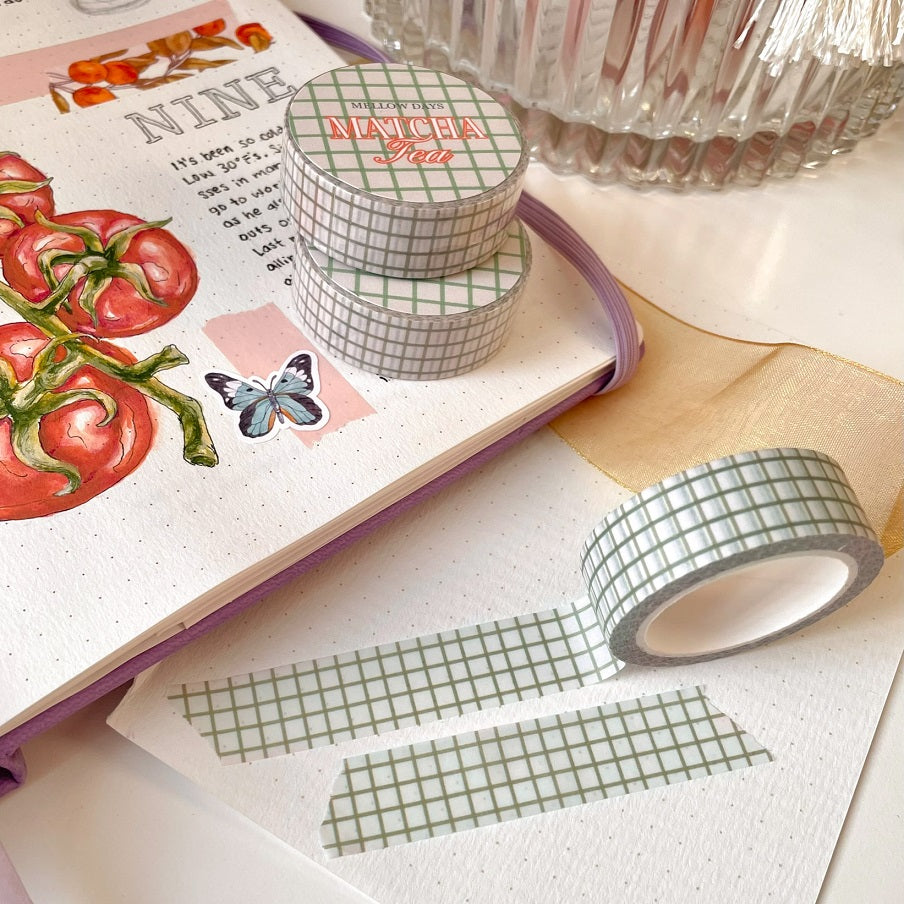 A roll of washi tape with a grid pattern in a sage green color, displayed on an open journal with bullet journal-style drawings and notes.